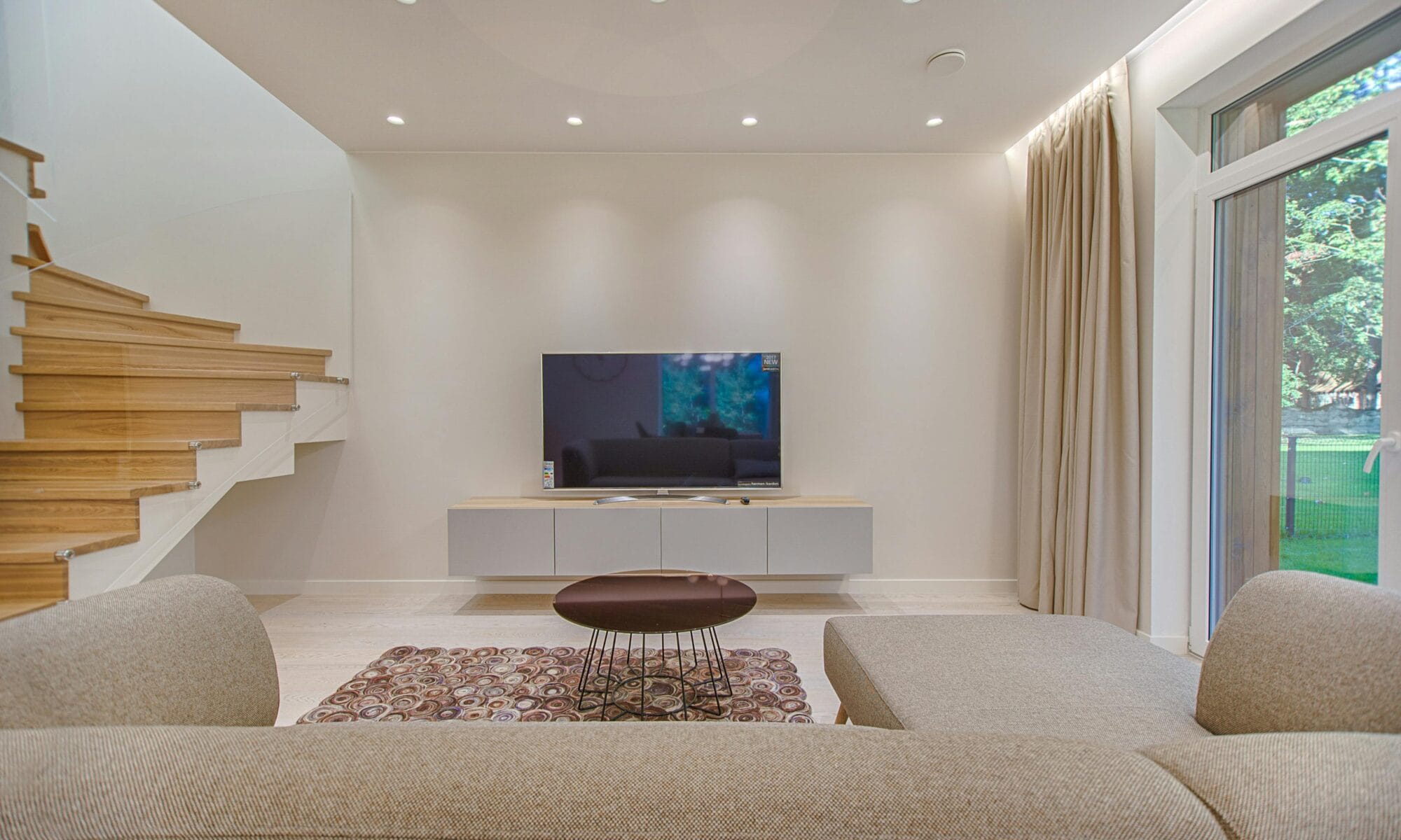 Minimalistisch ingerichte woonkamer met neutrale kleuren, voorzien van een zwevend tv-meubel, twee comfortabele grijze banken, en een unieke ronde koffietafel op een decoratief tapijt. De ruimte heeft grote ramen die uitkijken op een groene tuin en een moderne houten trap die naar de bovenverdieping leidt.